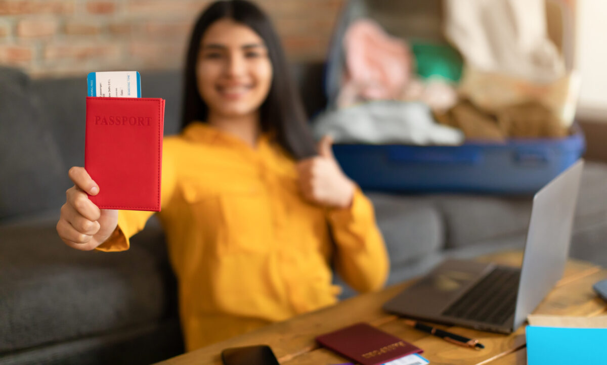 Smiling mexican lady showing passport with air tickets and gesturing thumb up, sitting with unpacked suitcase at home, selective focus. Emotions from travel, prepare to trip and vacation